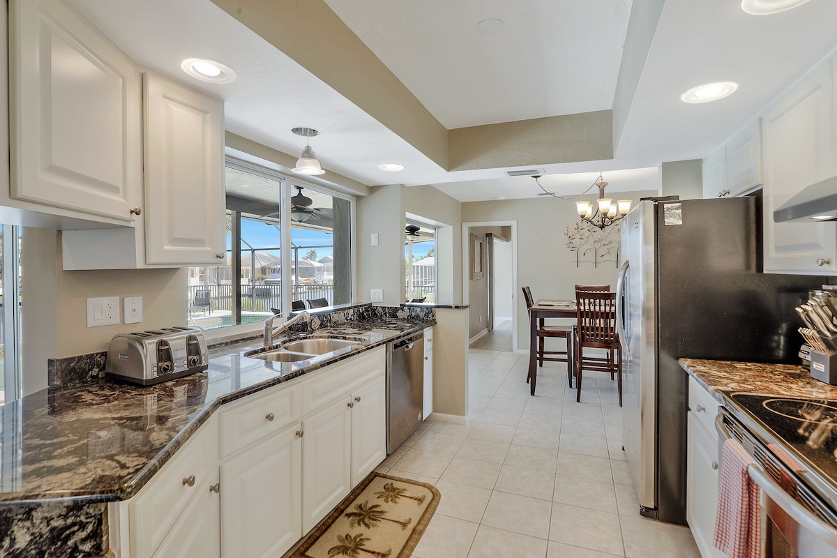 A modern kitchen with white cabinets, granite countertops, a stainless steel refrigerator, and a dining area with a chandelier.
