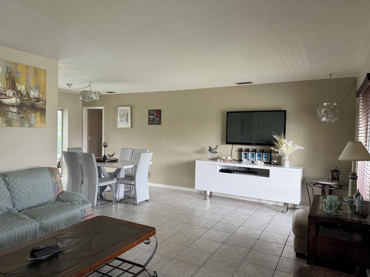 A living room with a sofa, a coffee table, a dining area with chairs, a white cabinet with decorative items, and a TV mounted on the wall.
