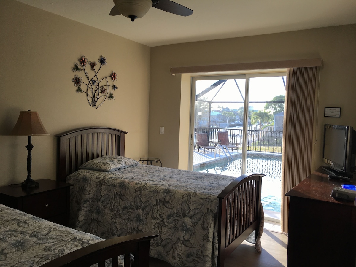 A bedroom with a bed, a desk with a TV, a lamp, and a window with a view of a pool and outdoor area.