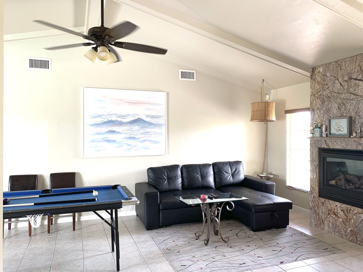 A living room with a pool table, a black leather sofa, a fireplace, and a ceiling fan.
