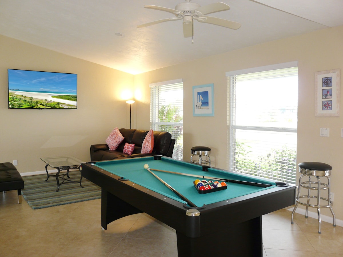 A pool table with a pool cue and balls, a sofa with patterned cushions, a framed picture on the wall, and a ceiling fan.