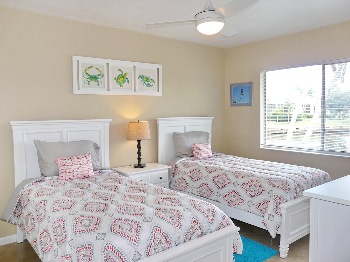 A bedroom with two beds, a nightstand with a lamp, a ceiling fan, and artwork on the wall.