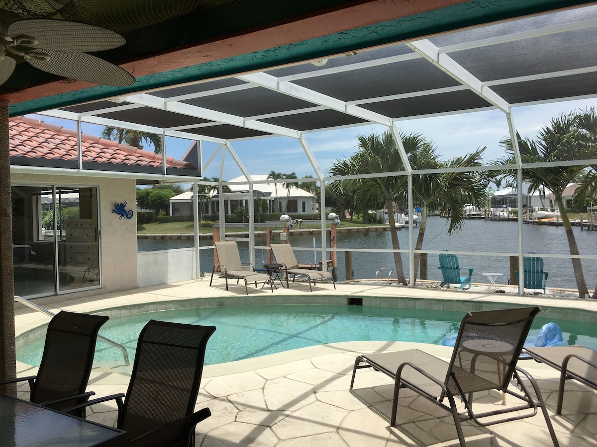 A pool area with chairs and a table, a covered patio, and a view of a body of water with palm trees.
