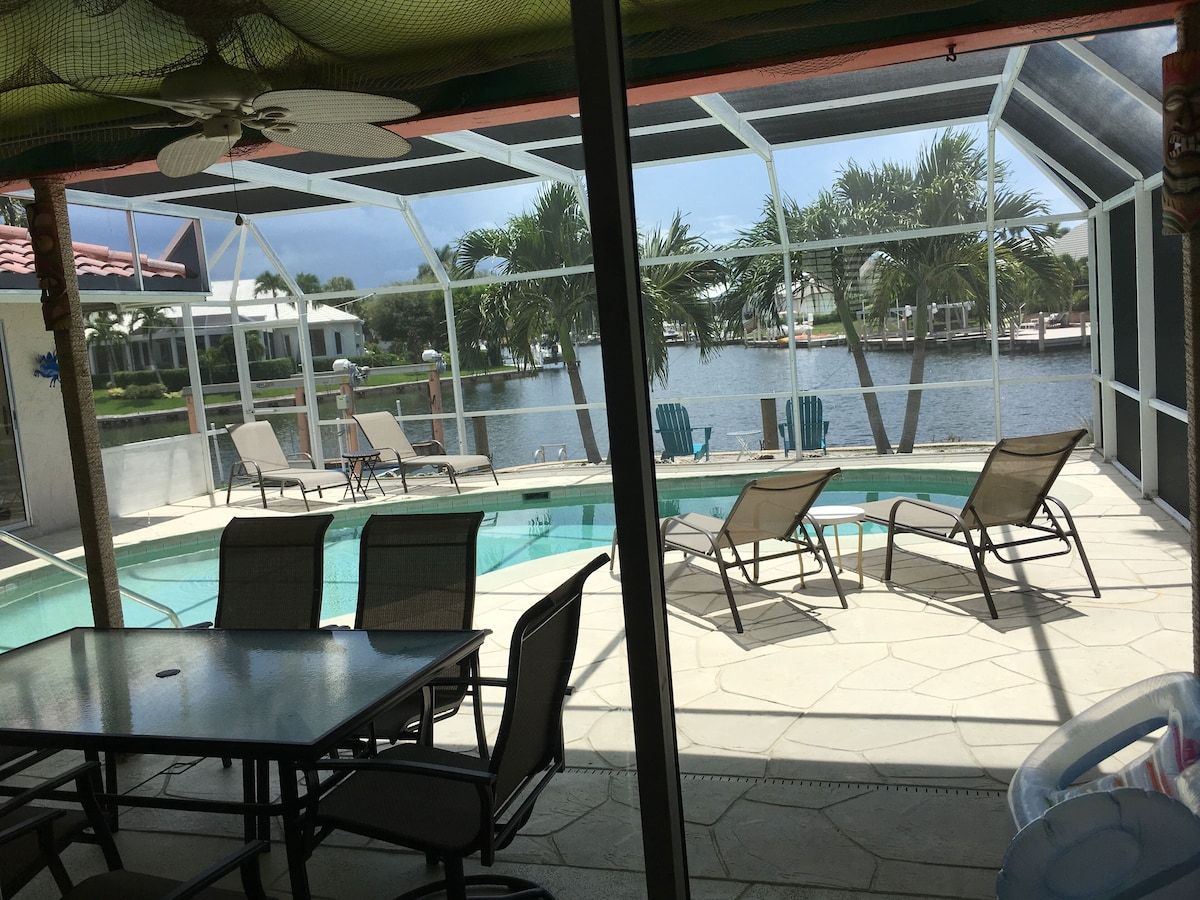 A poolside area with lounge chairs and a table, overlooking a body of water and greenery.