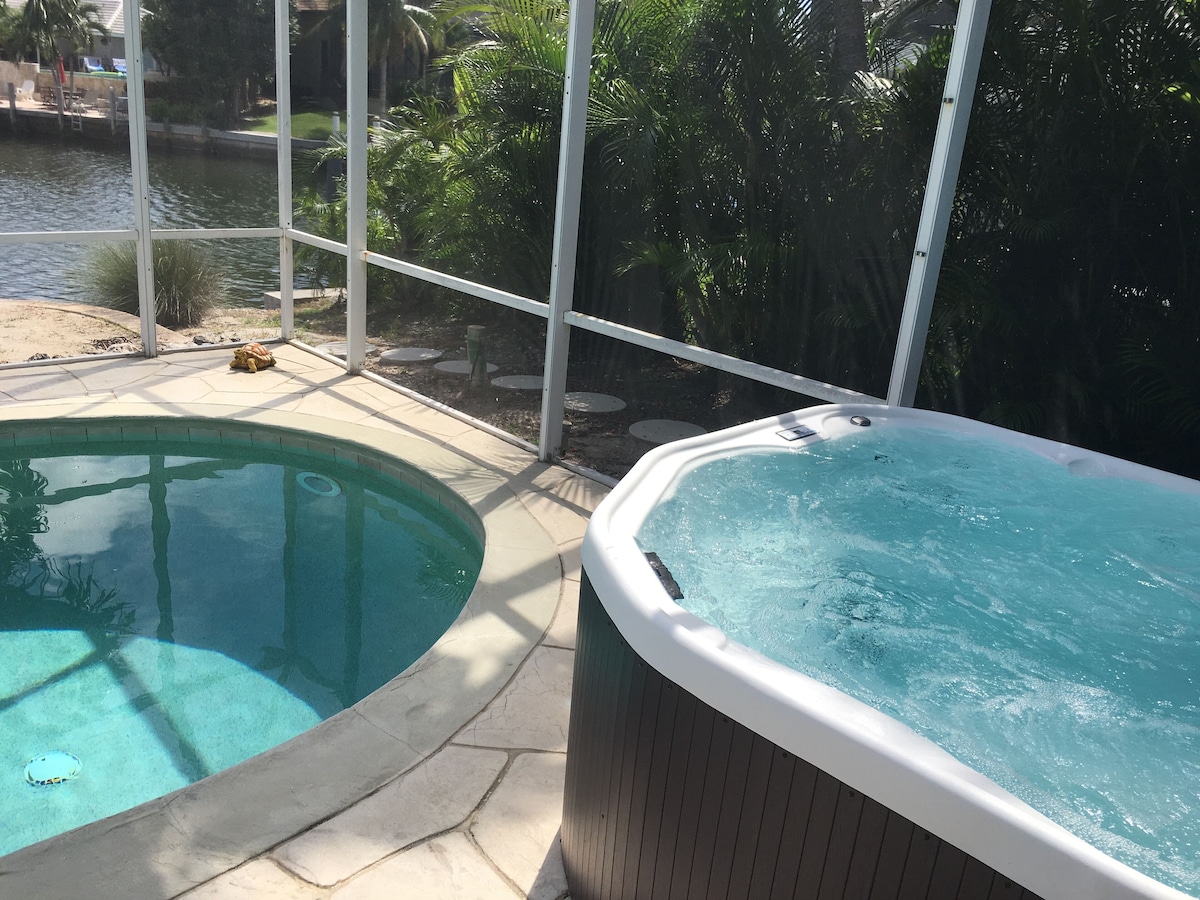 A swimming pool and a hot tub adjacent to a glass enclosure with a view of a body of water and greenery.