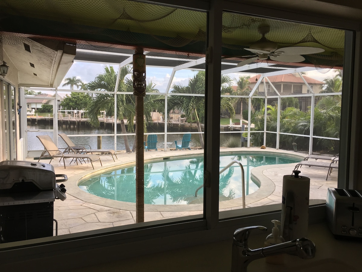 A pool area with lounge chairs, a grill, and a view of a water body and palm trees.