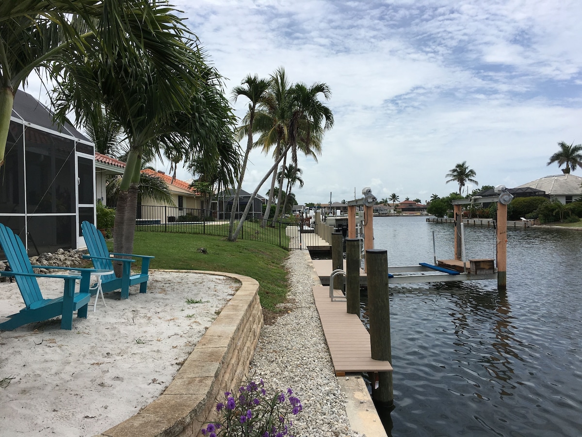 A waterfront property with a sandy patio, blue chairs, a wooden dock, and a view of the water.