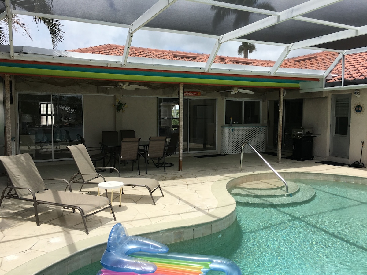 An outdoor pool area with lounge chairs, a covered patio, and a view of a red-tiled roof building.